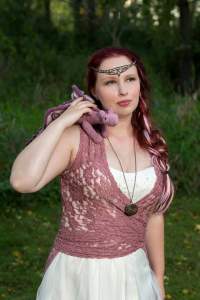 A young white woman with deep red hair looks wistfully off into the distance. She is wearing a pink lace vest over a white wedding dress, a jeweled headband, and a small, pink crocheted dragon on her shoulder. 