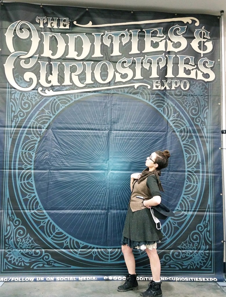 The writer, a brunette white woman with a short side cut & a bun, glasses, and multiple layered skirts, carrying a mask and a hat, looks up at a banner reading "The Oddities & Curiosities Expo." The backdrop is dark blue with an Art Deco circular design.