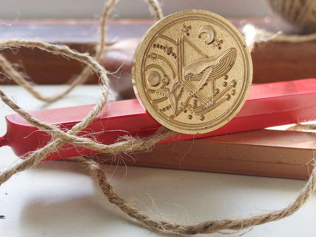 A metal seal with a whale swimming among stars, planets, a crescent moon, and three quadrilateral shapes faces the viewer. It is balanced on a stick of red sealing wax on top of a stick of metallic copper sealing wax. Hemp cord is draped over the pile.