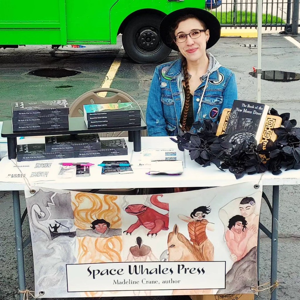 The writer, a medium-sized white woman with her hair in a braid, wears a jean jacket and a brimmed black hat. She sits behind a table stacked with books, with a banner with scenes from Beyond the Frost-Cold Sea hanging from the front.