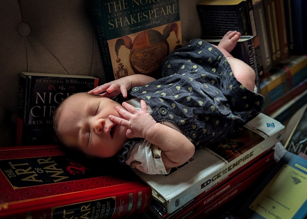 A baby wearing a black dress with gold hearts lies on a stack of books, including the Edge of the Empire RPG sourcebook, the D&D Monster Manual, Player's Handbook, and Dungeon Master's Guide, The Art of War, the Norton Shakespeare Anthology, The Golden Compass, Dune, The Night Circus, and Gulliver's Travels. She is turned toward the camera, eyes closed, her hands in her face. 